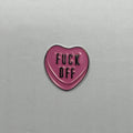 Pink heart-shaped metal sticker with 'FUCK OFF' text on a gray background