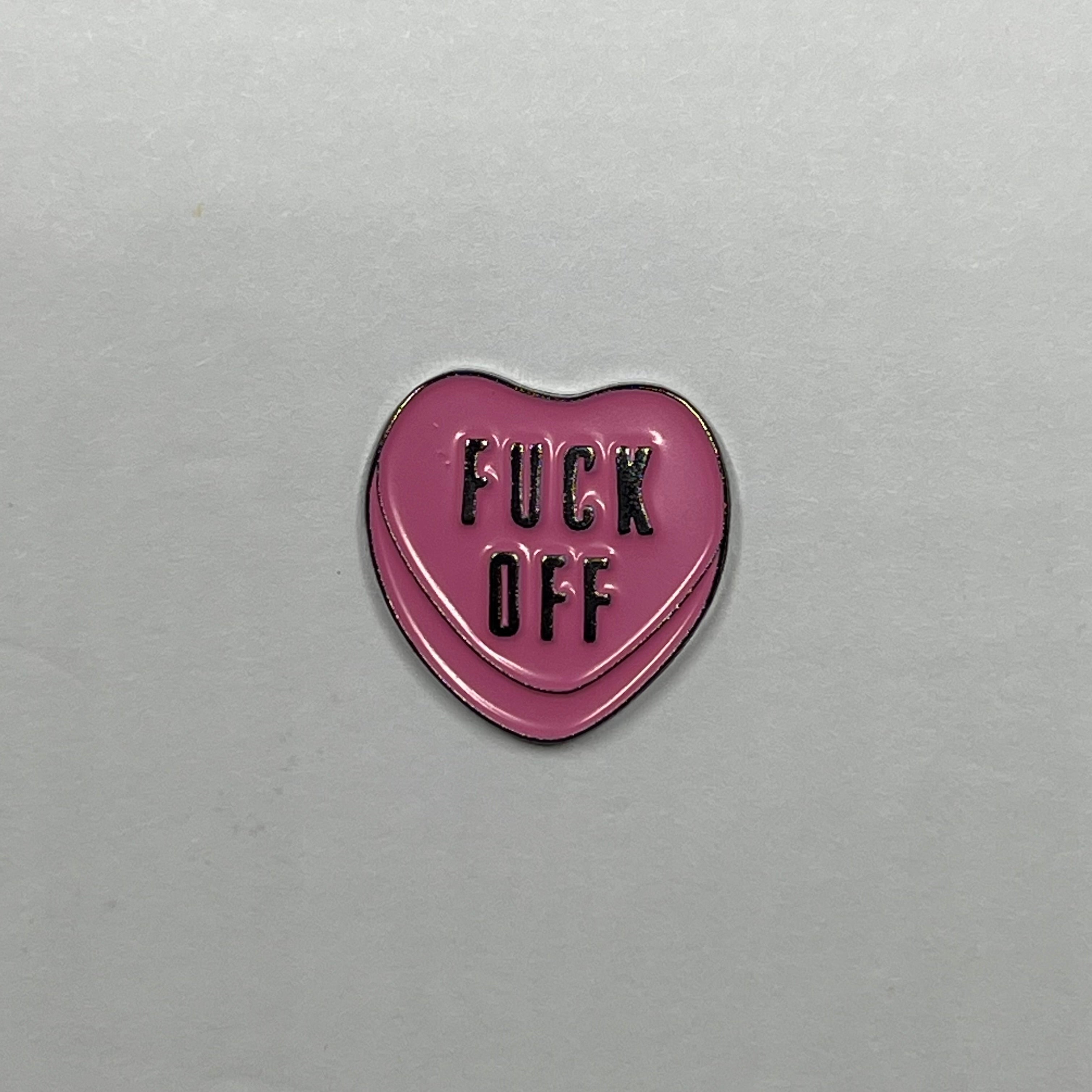 Pink heart-shaped metal sticker with 'FUCK OFF' text on a gray background